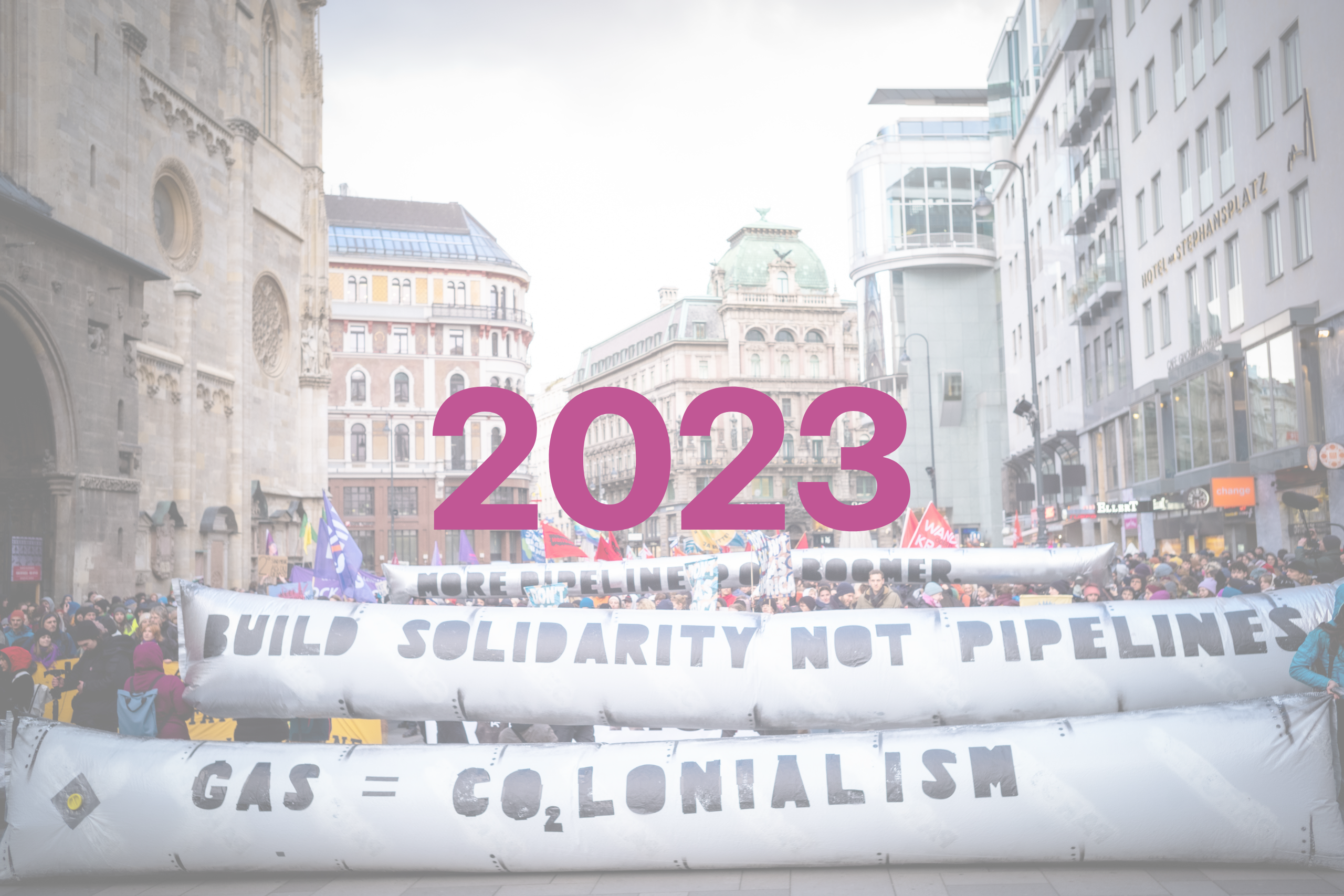 Man sieht die Zahl 2023, im Hintergrund ein Foto einer großen Demonstration. Die vordersten Reihen der Demonstrierenden halten zwei lange Banner übereinander, die aufblasbar sind und an riesige Rohre erinnern. Auf dem oberen Banner steht "build solidarity not pipelines", auf dem unteren Banner steht "gas = colonialism". Nach dem dem "CO" von "colonialism" steht noch die Zahl 2 - für CO2.